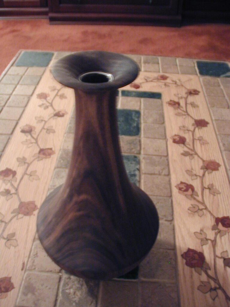 Tall Dark Wooden Vase with Flared Top and Narrow Neck Placed on a Decorative Tile Table